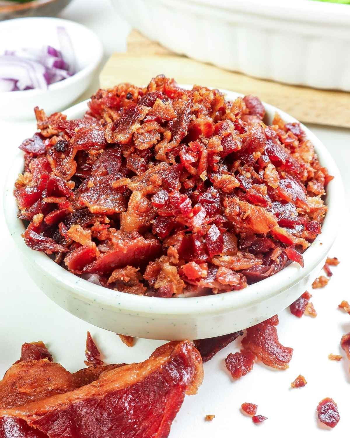 a bowl of bacon bits with a slice of cooked bacon on the side