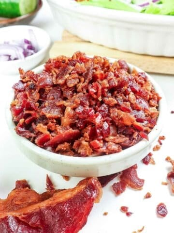 a bowl of bacon bits with a slice of cooked bacon on the side