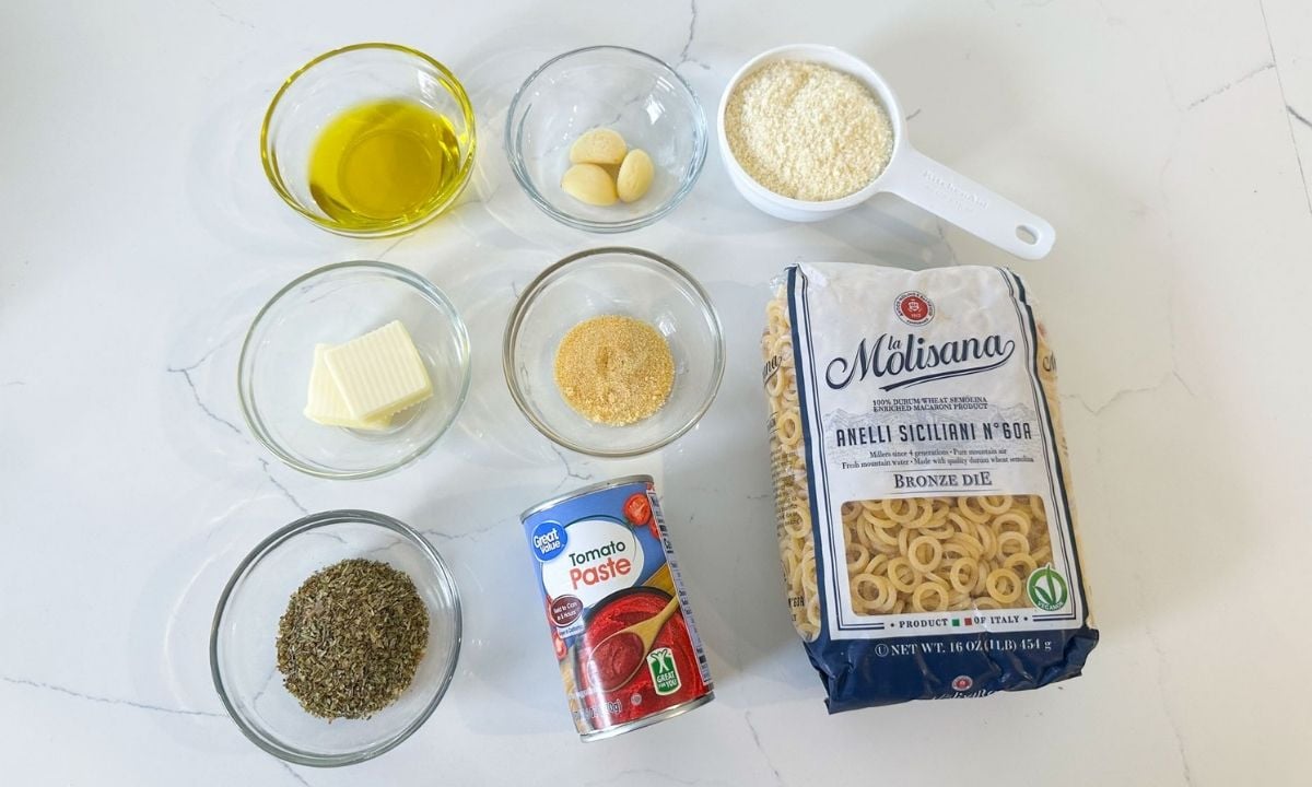 Ingredients for homemade SpaghettiOs, including anelli pasta, tomato paste, grated Parmesan cheese, olive oil, butter, garlic, Italian seasoning, and onion powder arranged on a white countertop.