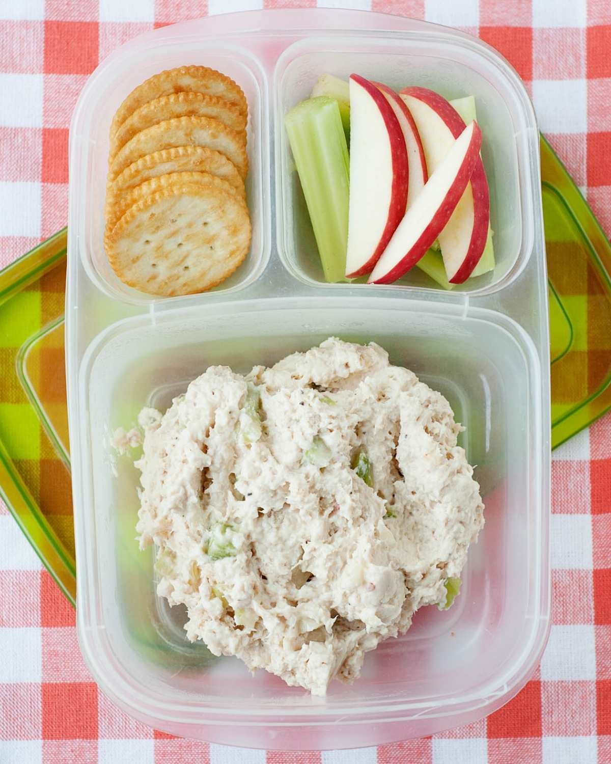 a lunchbox with crackers, sliced apples and celery, and chicken salad in the biggest compartment