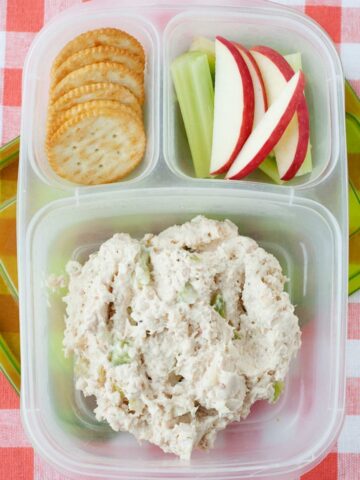 a lunchbox with crackers, sliced apples and celery, and chicken salad in the biggest compartment