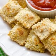 a white dish with baked ravioli and a small bowl of marinara sauce