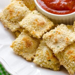 a white dish with baked ravioli and a small bowl of marinara sauce