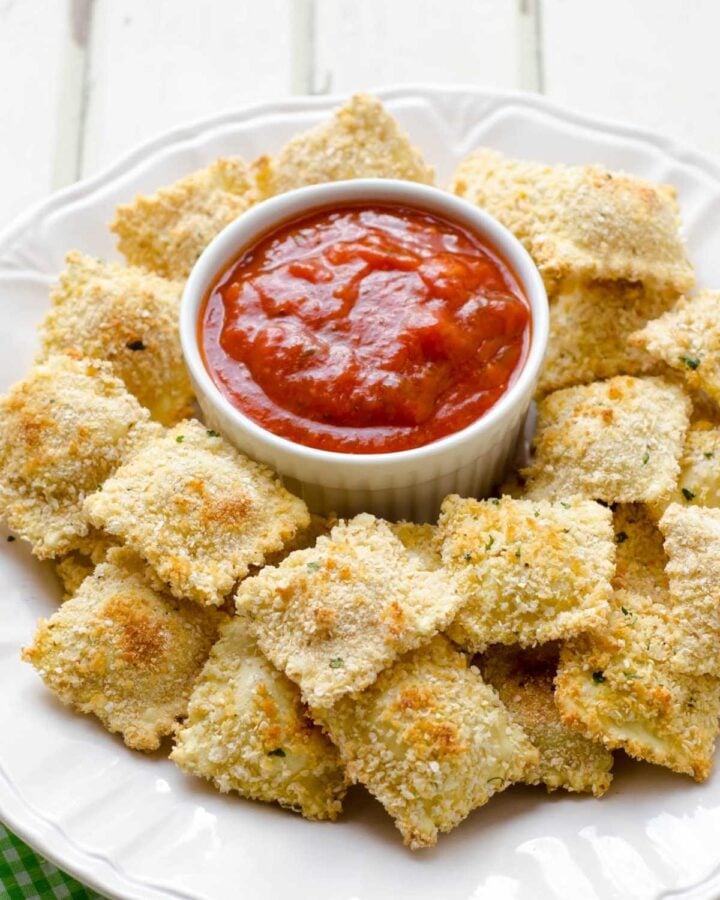 a white dish with baked ravioli surronding a small bowl of marinara sauce