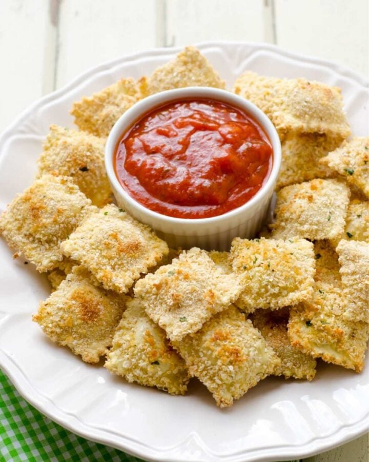 a white dish with baked ravioli surronding a small bowl of marinara sauce