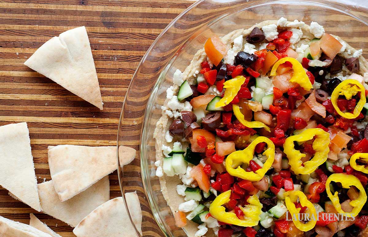 layered greek hummus dip in a glass bowl on a wooden board