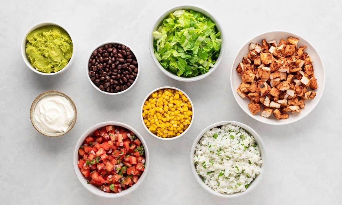small white bowls individually filled with guacamole, sour cream, black beans, pico de gallo, corn, cilantro lime rice, cooked chicken cubes, and lettuce