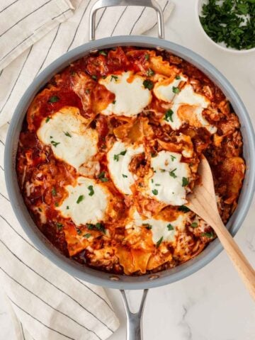 overhead view of a skillet with lasagna made with lasagna noodles, tomato sauce, meat, and cheeses using a wooden serving spoon to serve