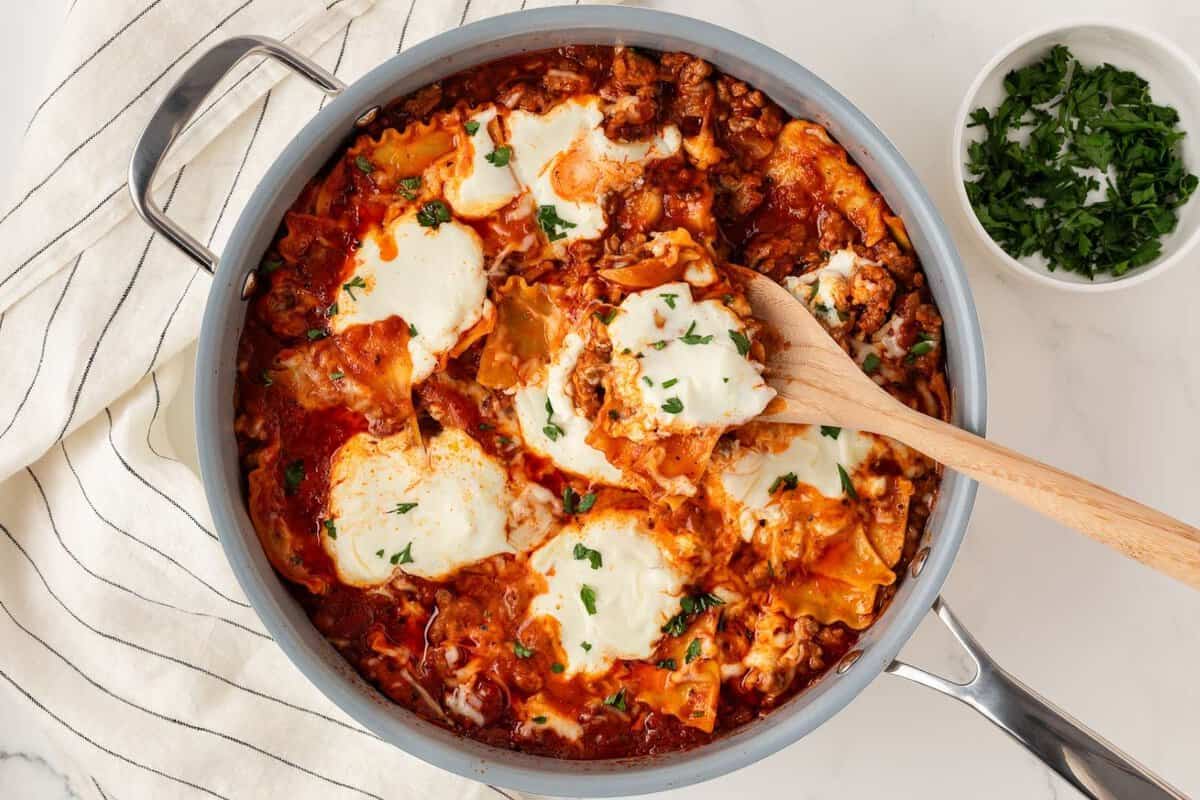 overhead view of a skillet with lasagna made with lasagna noodles, tomato sauce, meat, and cheeses using a wooden serving spoon to serve