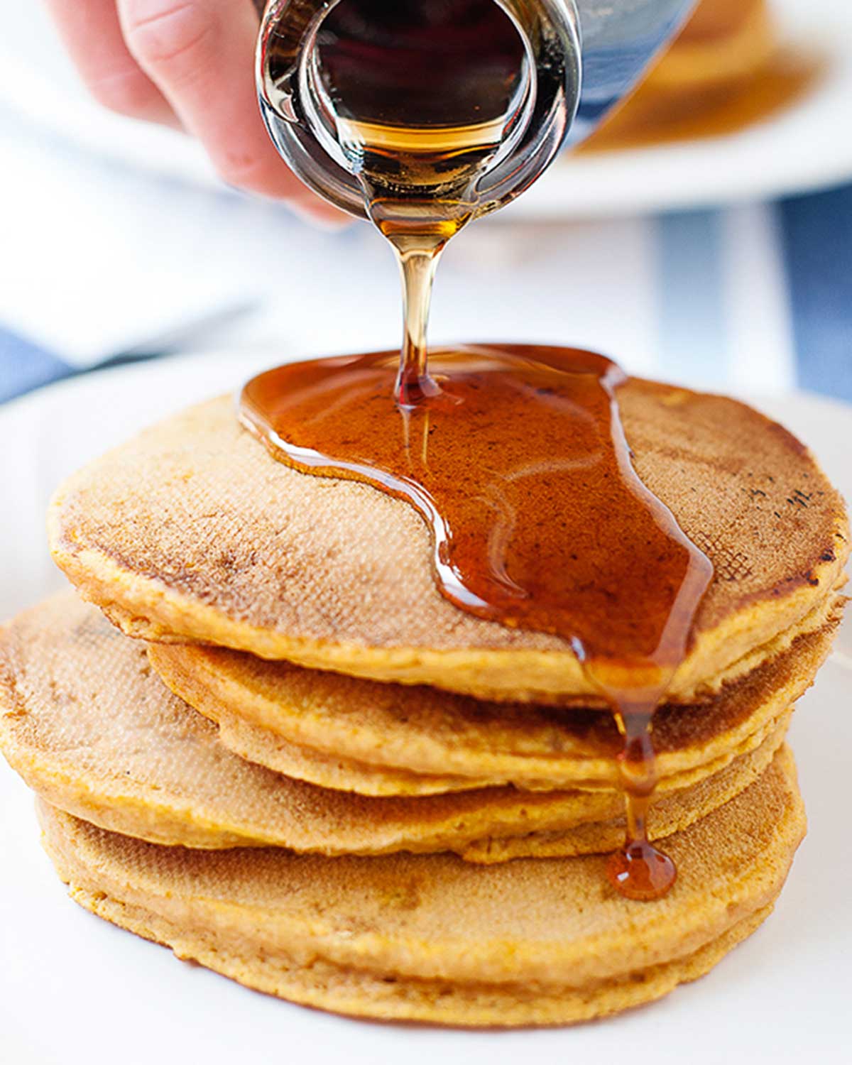 pumpkin pancakes on a plate stacked high and maple syrup being drizzled over the top dripping on the sides
