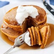 stack of pumpkin pancakes topped with whipped cream and syrup on a plate. A slice is cut and pieces through a fork