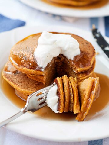 stack of pumpkin pancakes topped with whipped cream and syrup on a plate. A slice is cut and pieces through a fork