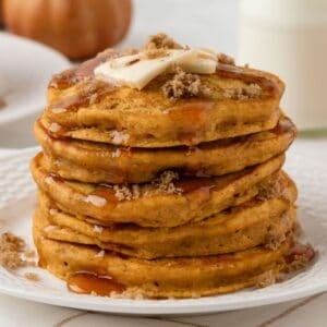 tall and fluffy stack of pumpkin pancakes on a plate topped with butter, brown sugar, cinnamon, and pancake syrup