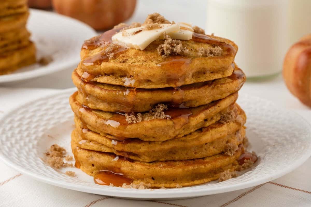 tall and fluffy stack of pumpkin pancakes on a plate topped with butter, brown sugar, cinnamon, and pancake syrup