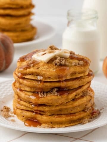 tall and fluffy stack of pumpkin pancakes on a plate topped with butter, brown sugar, cinnamon, and pancake syrup