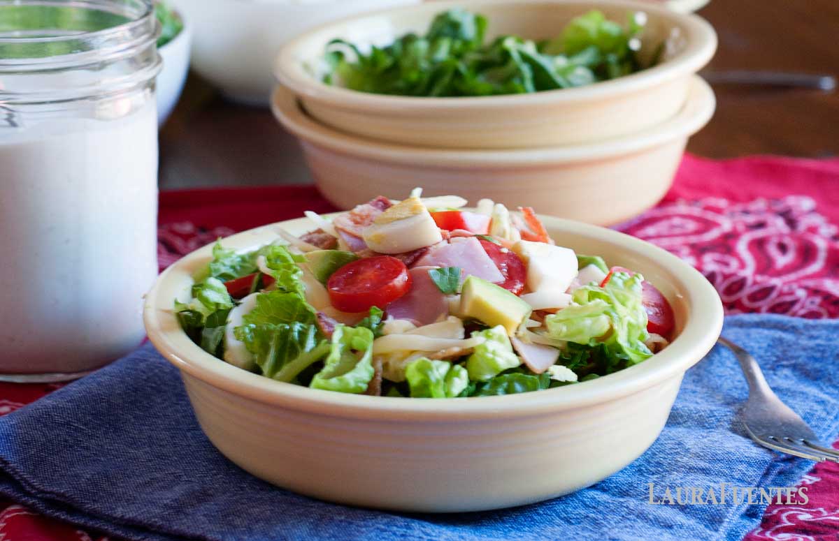 a bowl of cobb salad with the ingredients of cobb salad bar in the background