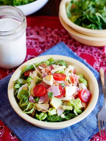 a bowl of cobb salad with ham, cheese, greens, hard boiled egg, and tomato, next to a jar of dressing and a bowl of greens