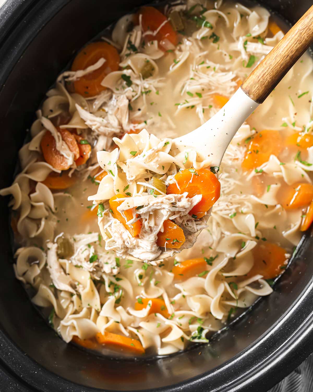 overhead view of chicken noodle soup inside a slow cooker being lifted in a ladle made with rotisserie chicken, egg noodles, carrots, celery, and a rich broth
