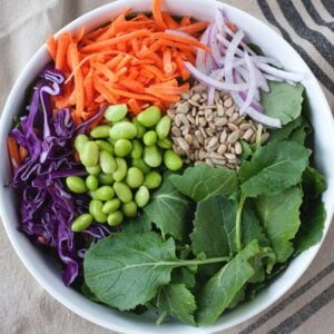 top view of a white bowl with edamame, kale, red cabbage, shredded carrot, onion and seeds