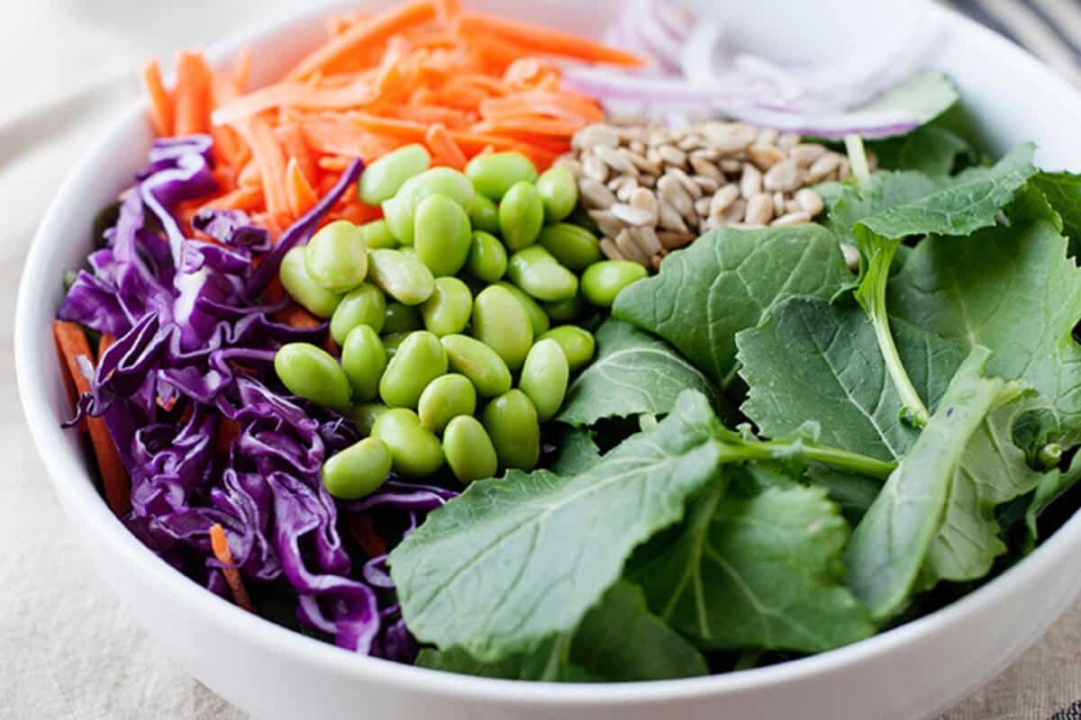top view of a white bowl with a salad made with edamame, kale, red cabbage, shredded carrot, onion and seeds