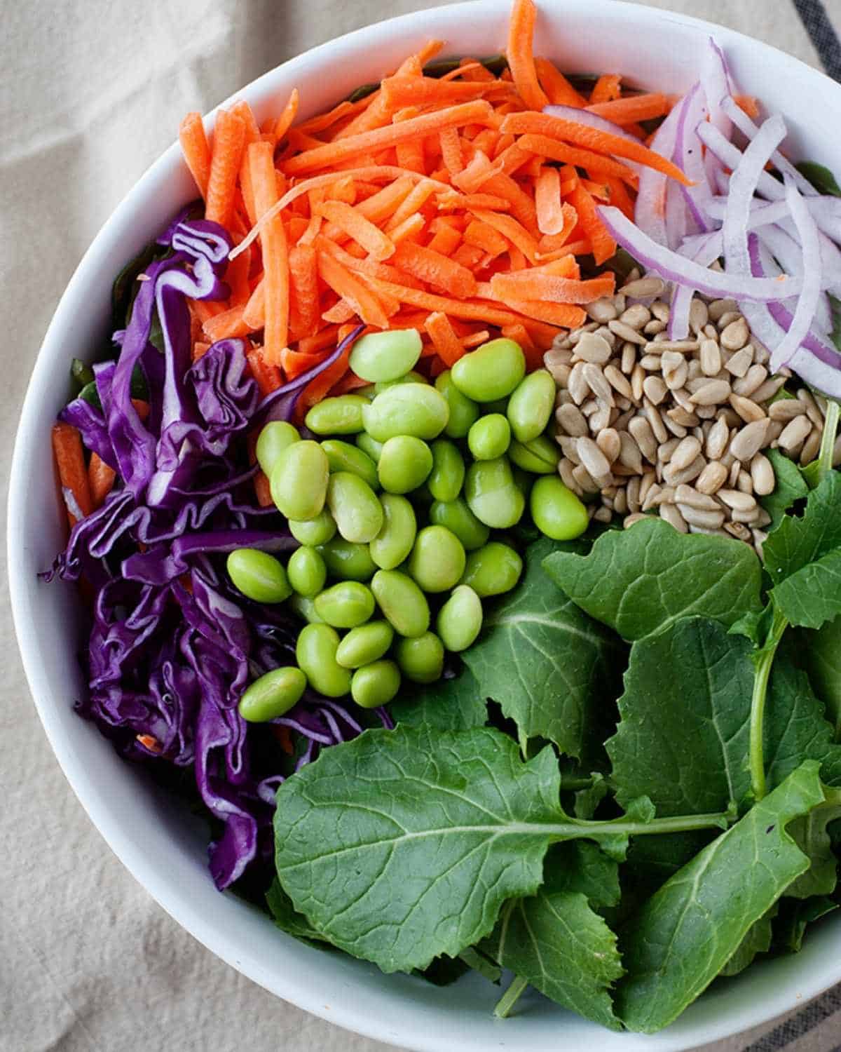 top view of a white bowl with edamame, kale, red cabbage, shredded carrot, onion and seeds