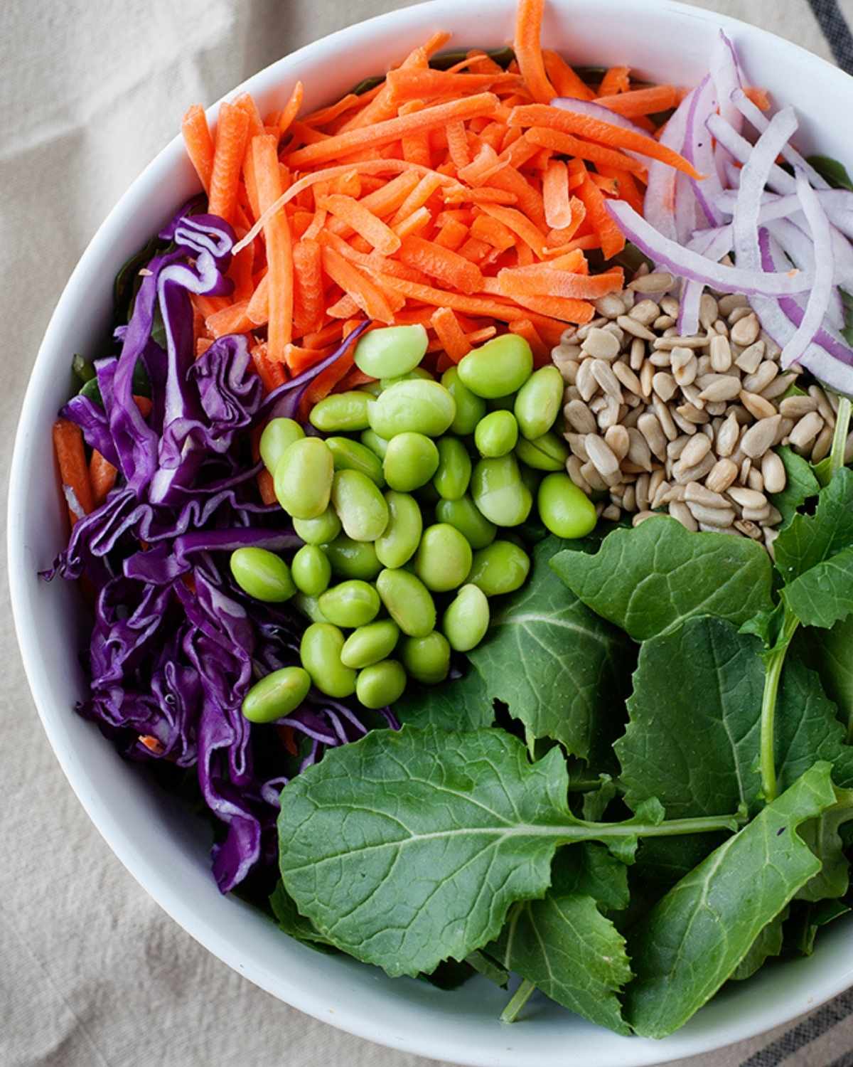 top view of a bowl with edamame, kale, red cabbage, shredded carrot, onion and seeds