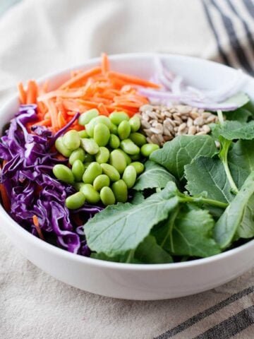 a white bowl with edamame, kale, red cabbage, shredded carrot, onion and seeds
