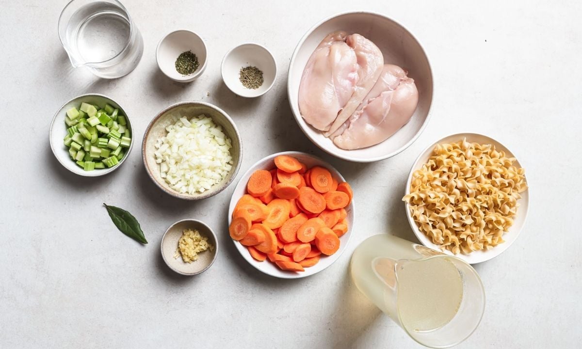 Ingredients for slow cooker chicken noodle soup, including chicken breasts, egg noodles, carrots, celery, onion, garlic, chicken broth, water, herbs, and a bay leaf arranged on a white countertop.