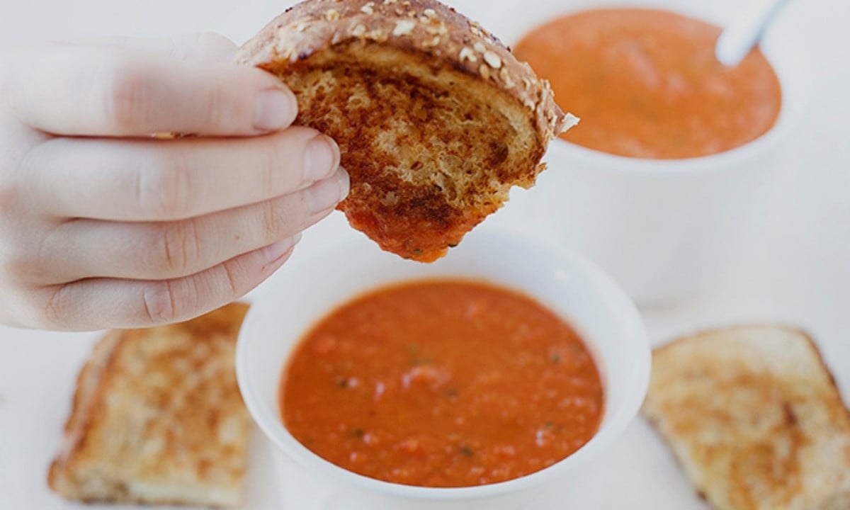 kid's hand dipping grilled cheese in a small bowl of tomato soup