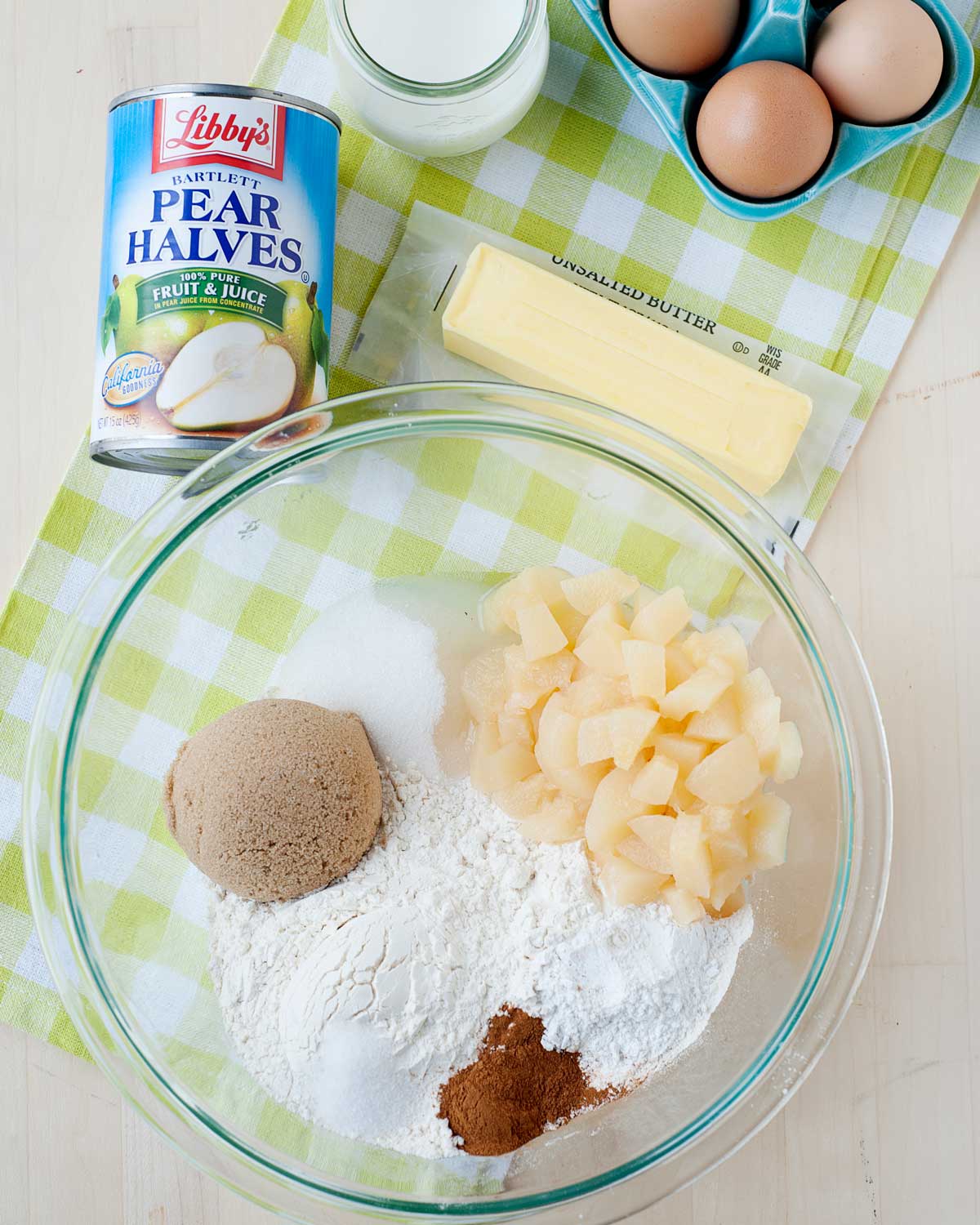 overhead view of a bowl with muffin ingredients (flour, sugar, cinnamon, baking powder) and chopped canned pears. there is a can of pears on the top, a stick of room temperature butter, milk, and eggs