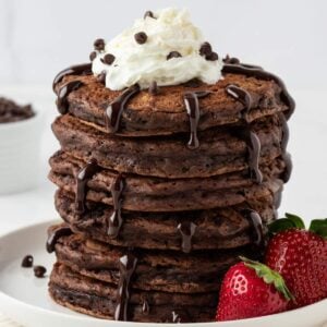 stack of chocolate pancakes with chocolate chips topped with whipped cream and chocolate chips and a drizzle of chocolate syrup on a plate with strawberries on the side.