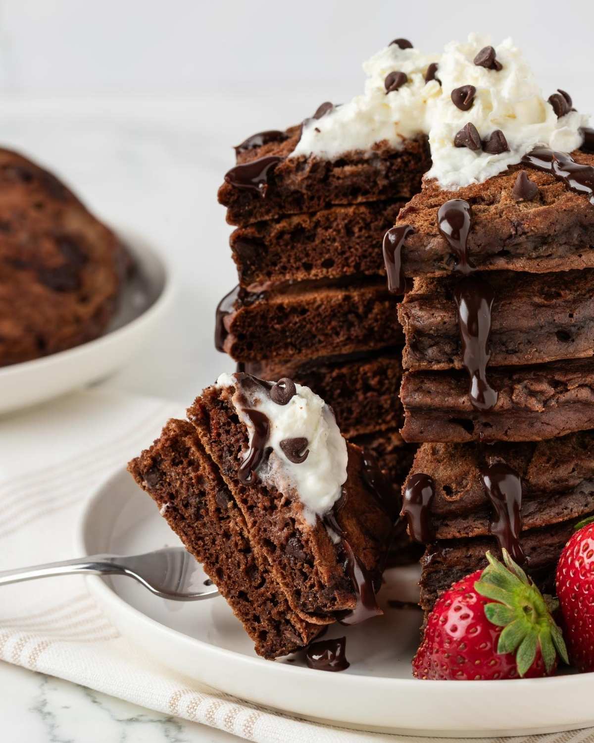 stack of chocolate pancakes with chocolate chips topped with whipped cream and chocolate chips and a drizzle of chocolate syrup on a plate with strawberries on the side.