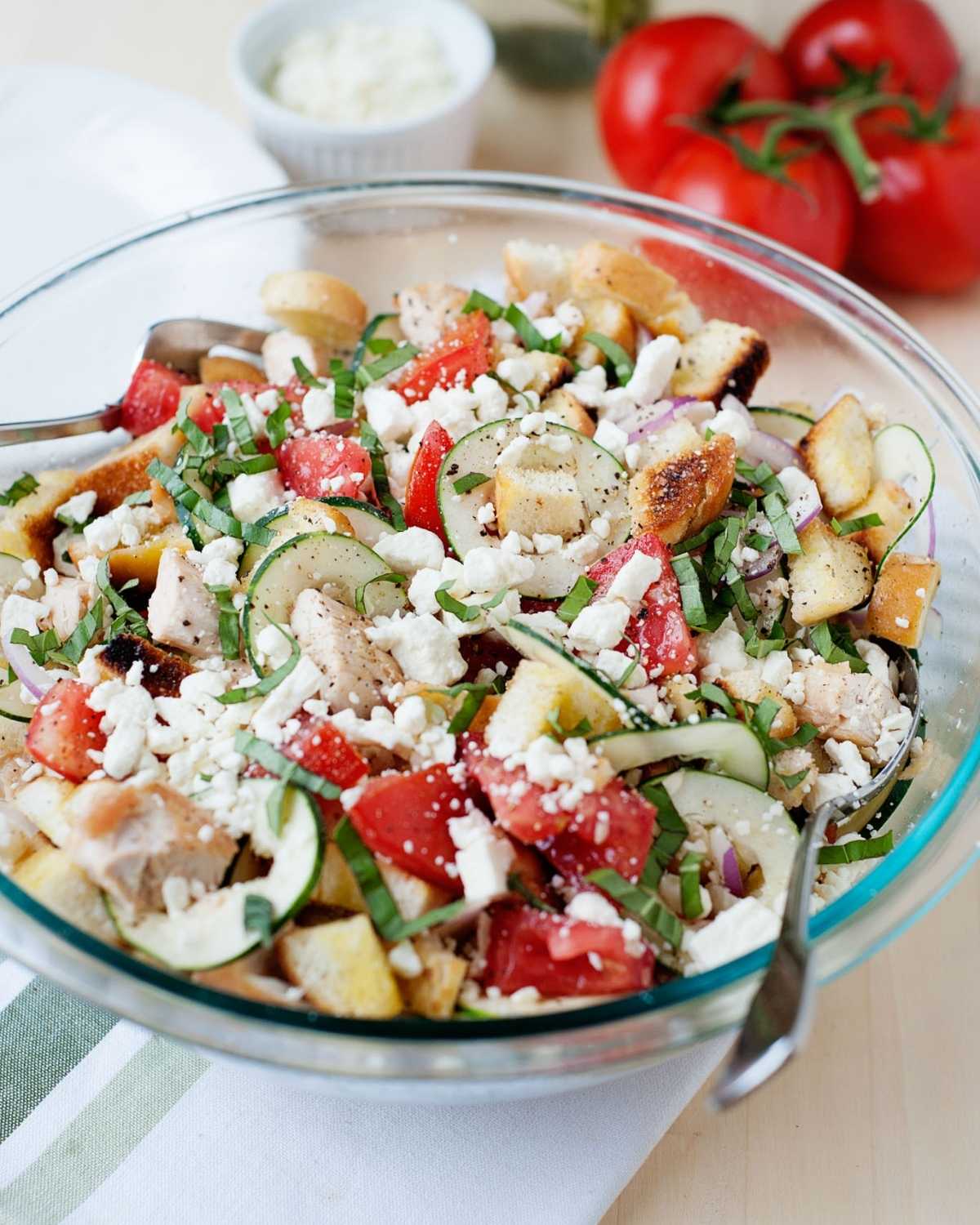 a bowl of panzanella salad with cucumber, tomato and crumbled cheese, and some tomatoes on the back