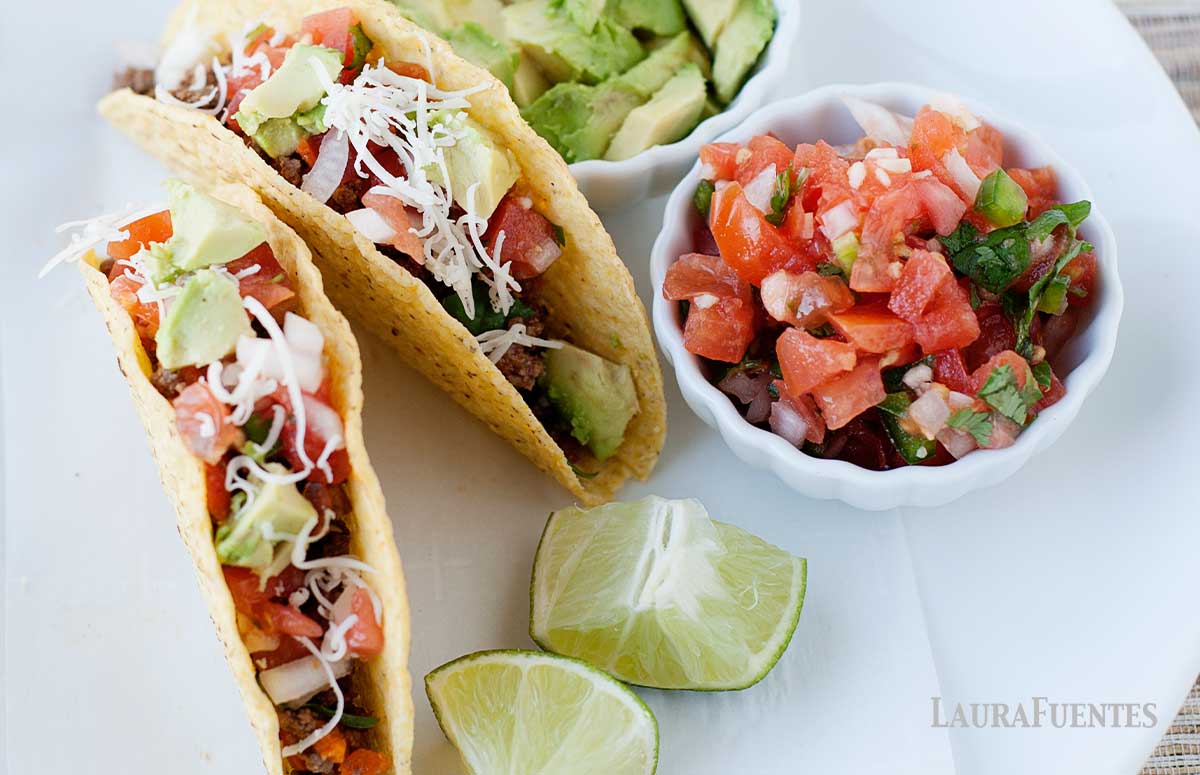 overhead view of kale tacos on a plate with limes, pico de gallo and sliced avocados on the slide