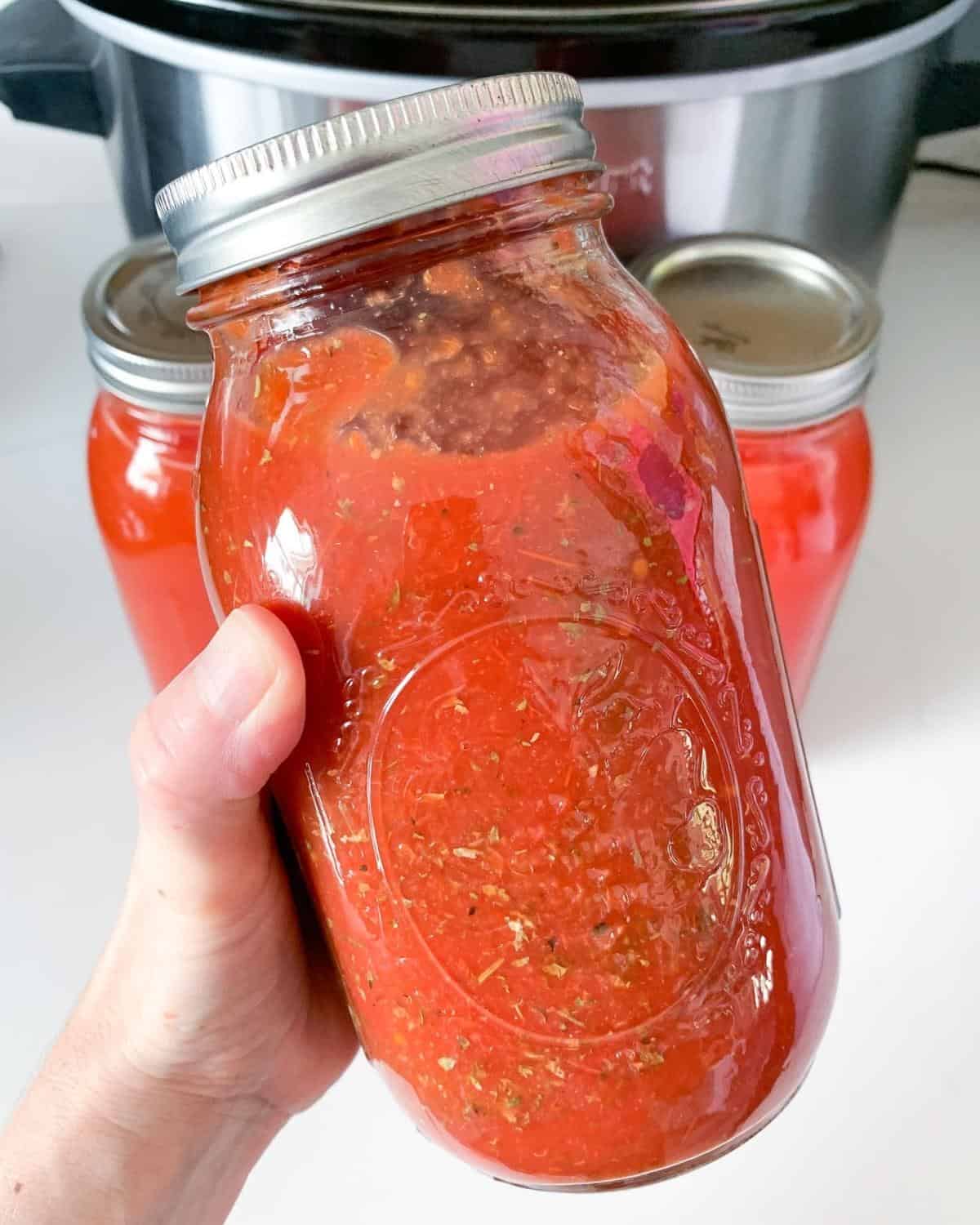 a hand holding a jar of tomato sauce, two more jars behind, and a slow cooker in the back