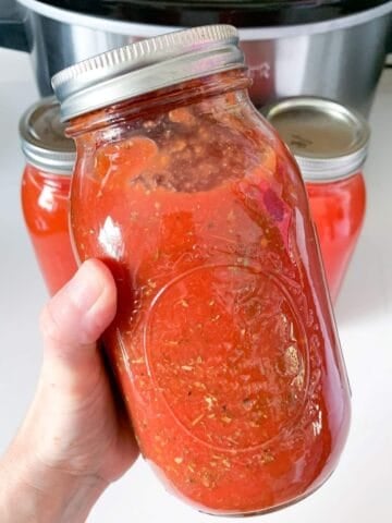 a hand holding a jar of tomato sauce, two more jars behind, and a slow cooker in the back