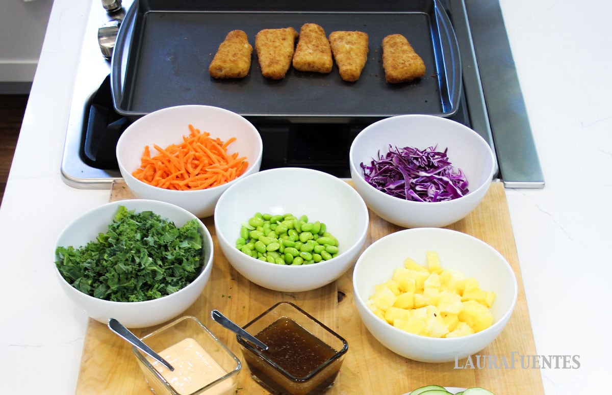 baking sheet with breaded fish and five bowls with shredded carrot, cabbage, kale, edamame and pineapple chunks