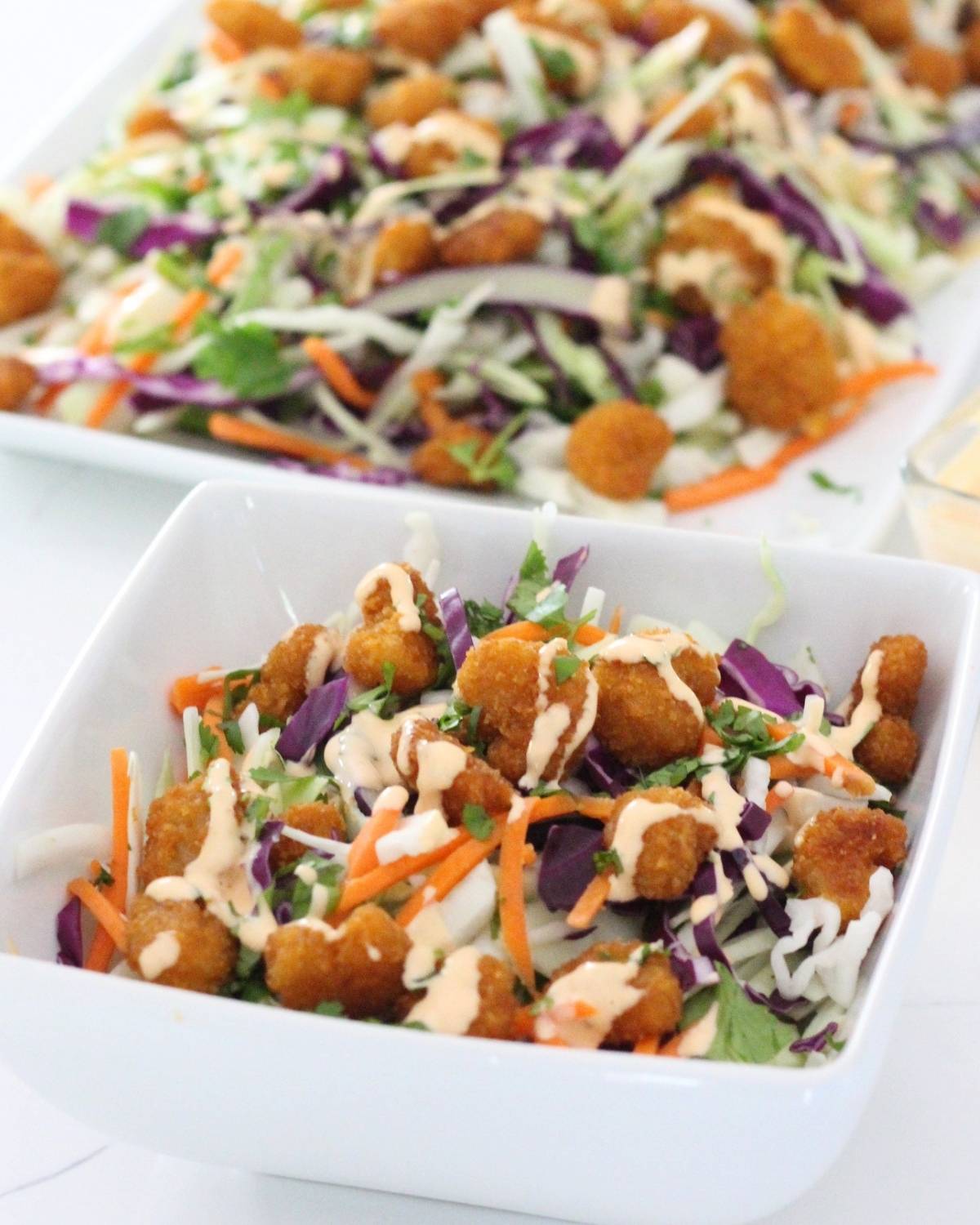 a bowl with popcorn shrimp salad with dressing and more salad behind on a platter
