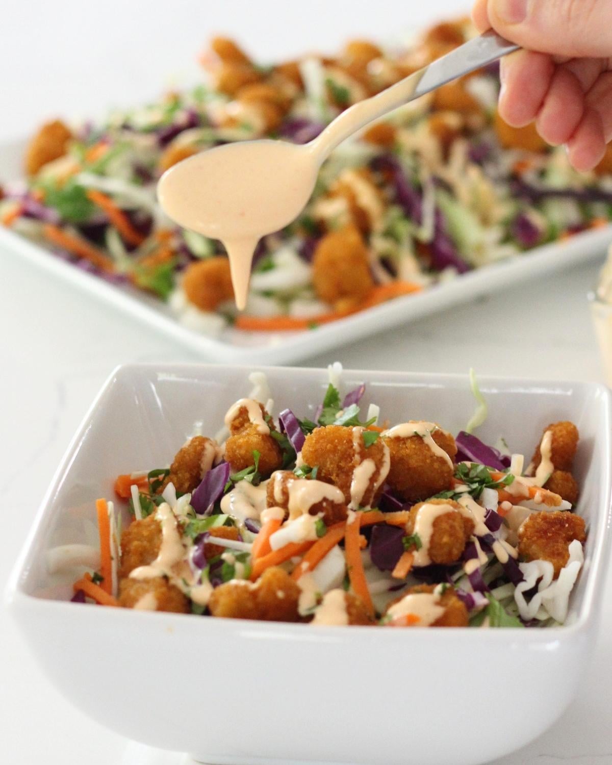 a hnd pouring dressing over a bowl of popcorn shrimp salad and more salad behind on a platter