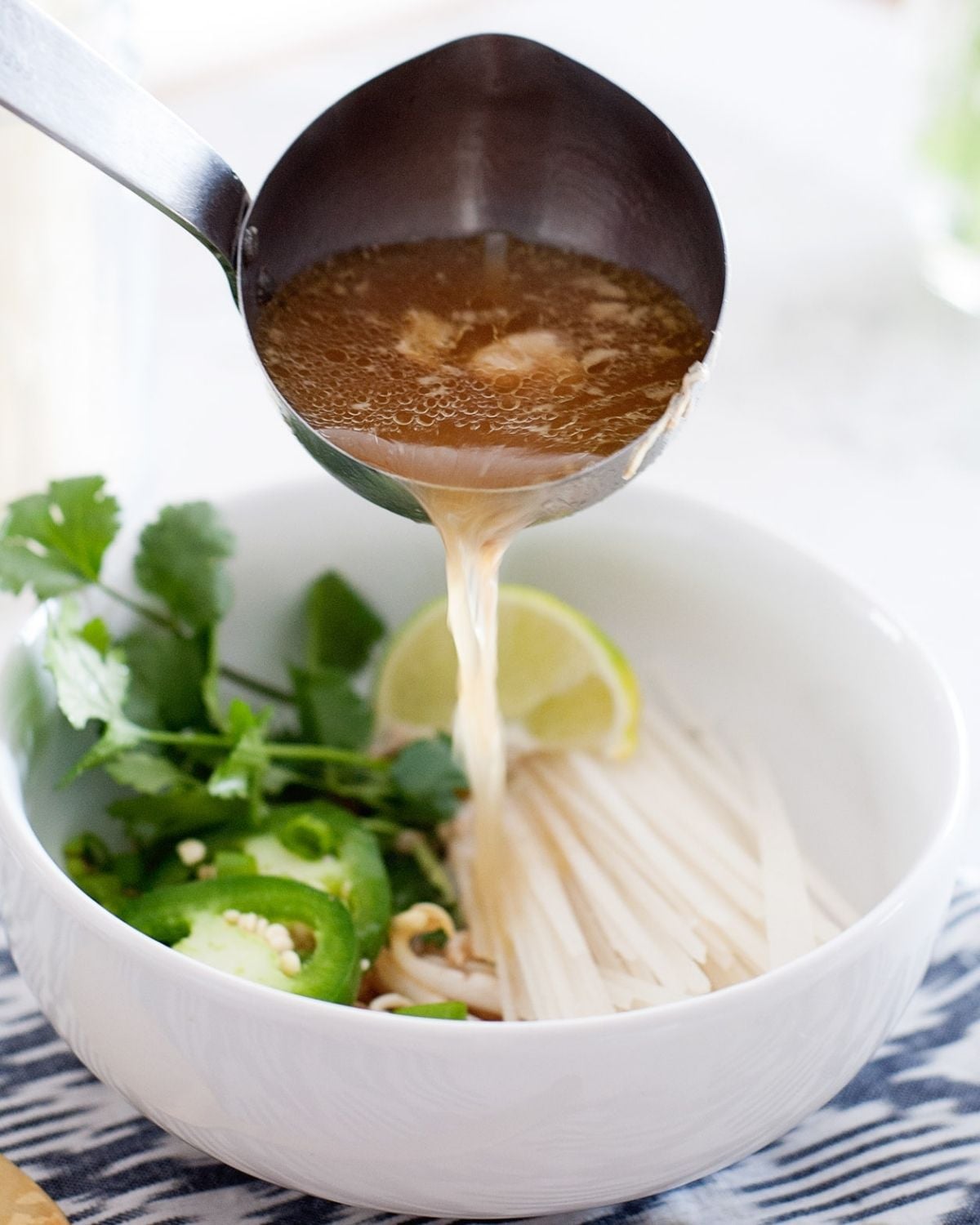 a soup spoon pouring broth over a bowl with rice noodles, jalapeños, sliced jalapñeo and a lime wedge
