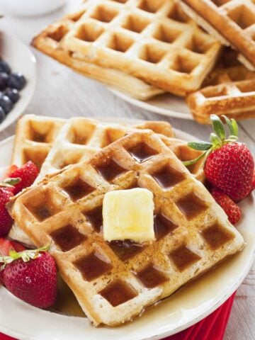 two buttermilk waffles on a plate topped with some butter and syrup with strawberries on the side. more buttermilk waffles in the background