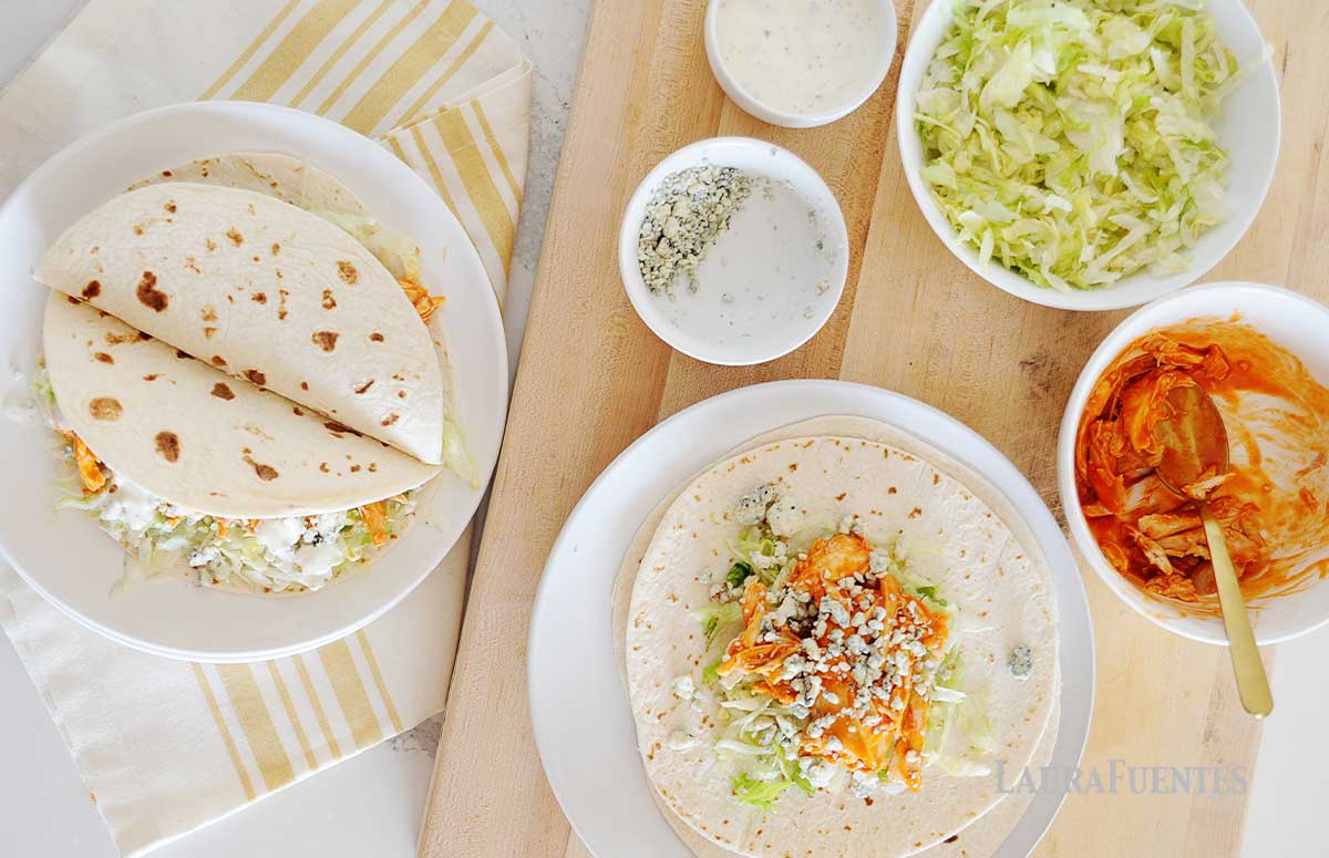 slow cooker buffalo chicken tacos on a kitchen counter, one of them is in the making with the ingredients on the side