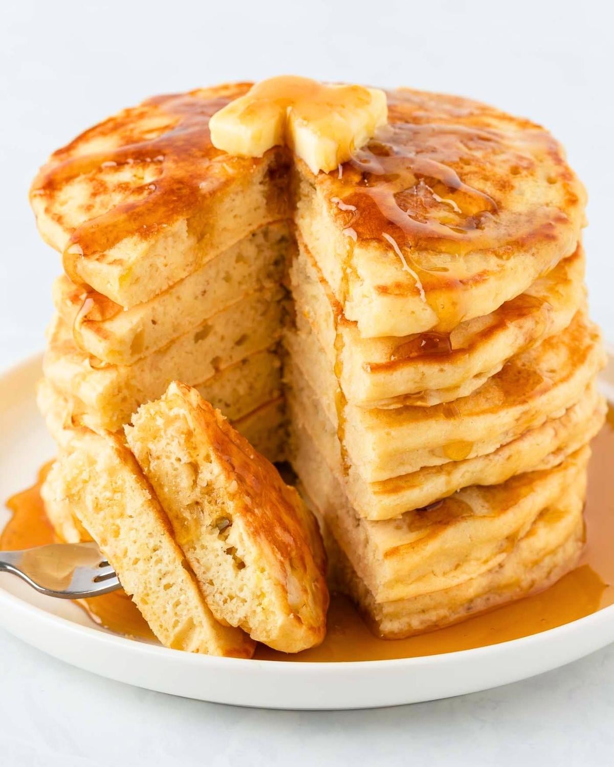 Stack of homemade pancakes on a plate topped with butter and syrup. they've been sliced to show their fluffy texture