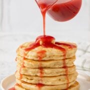 jar of strawberry syrup pouring over a large stack of pancakes