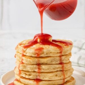 jar of strawberry syrup pouring over a large stack of pancakes