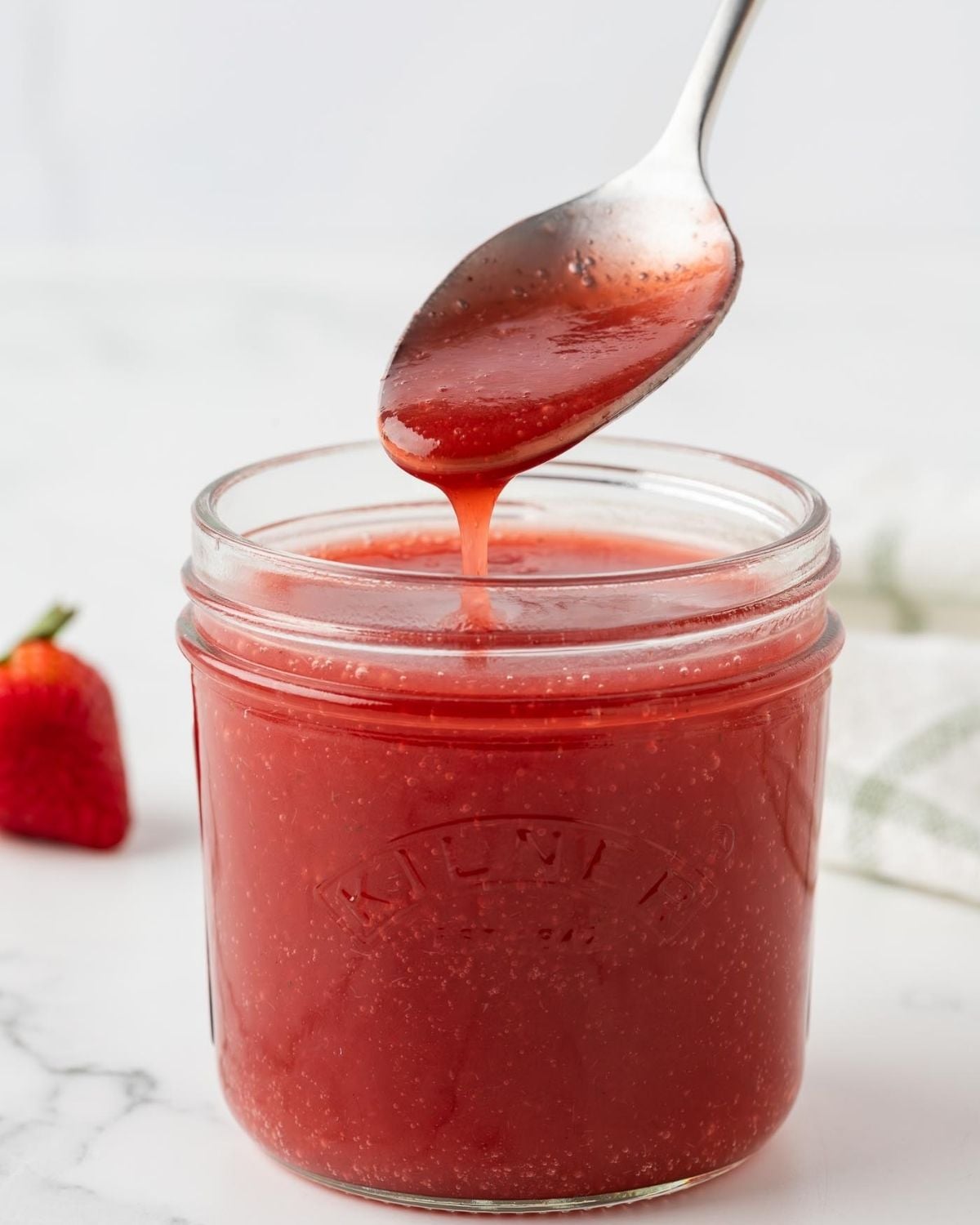 homemade strawberry syrup in a jar. a spoon pouring some to show thickness/texture
