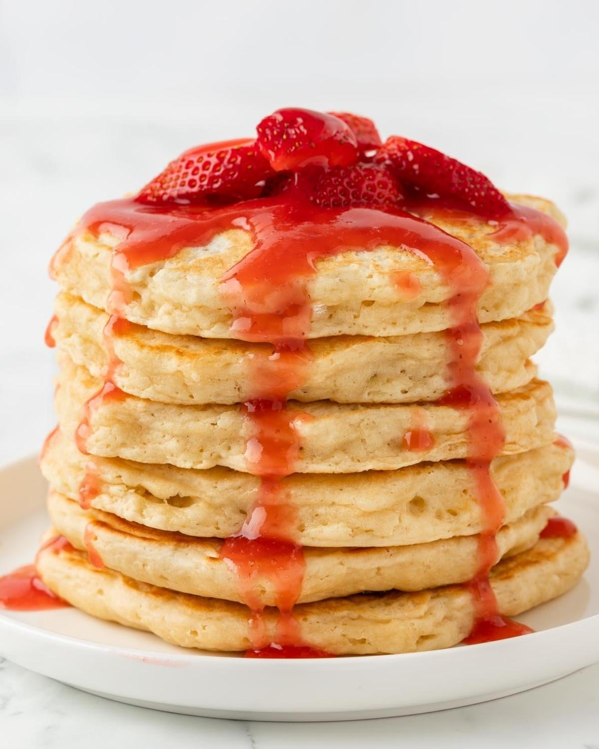 front view of a fluffy pancake stack topped with a few sliced strawberries and strawberry syrup falling down the sides