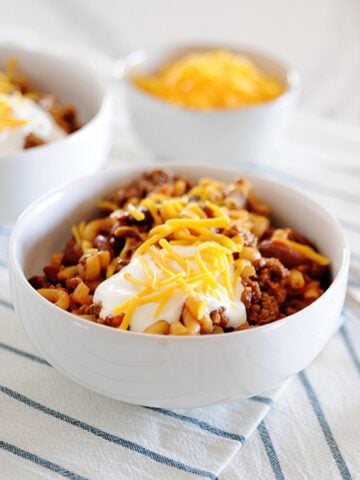 a bowl of chili mac topped with sour cream and cheese, and a small bowl of cheese in the background