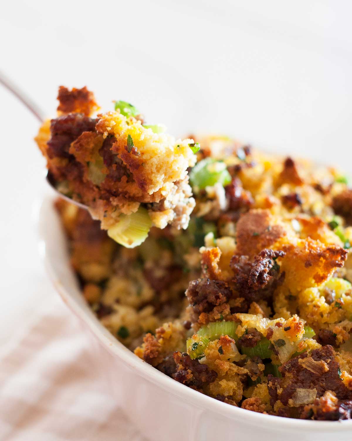close up view of a serving spoon lifting the cornbread dressing with sausage inside a white baking dish