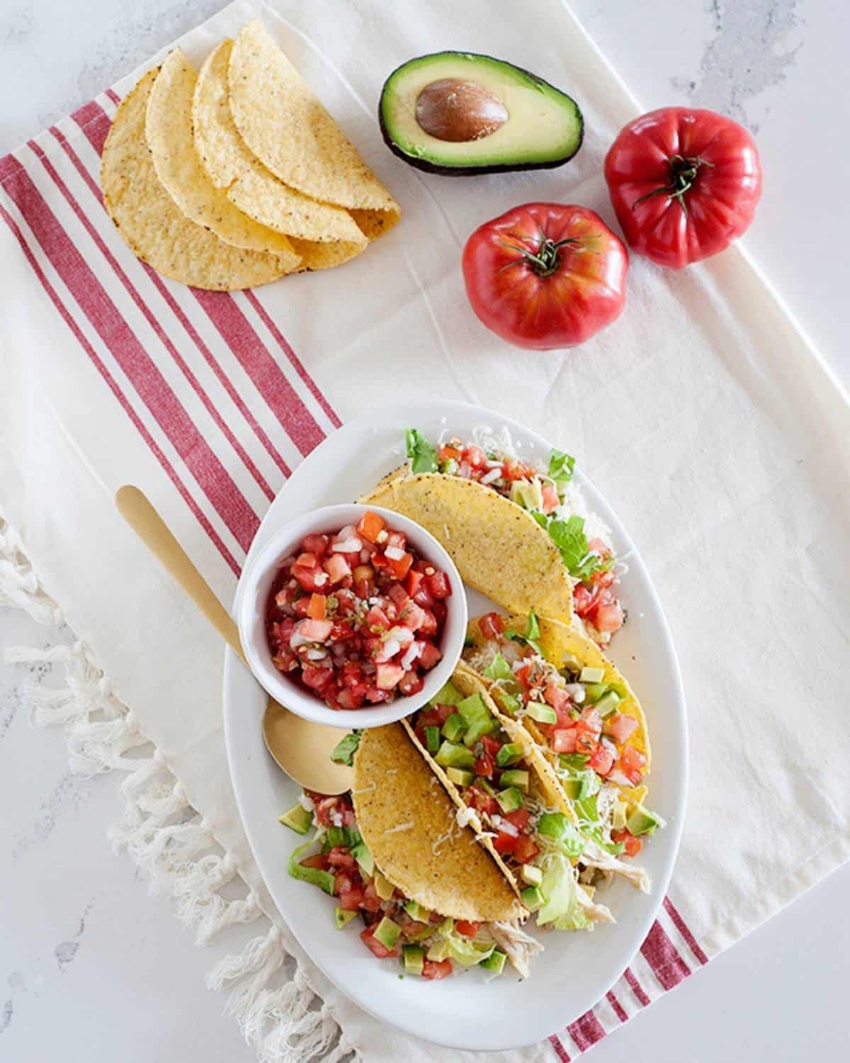 overhead view of Slow Cooker Salsa Verde Chicken Tacos on a white plate and a small bowl of pico, de gallo on the side, plus avocado, tomato and tortillas